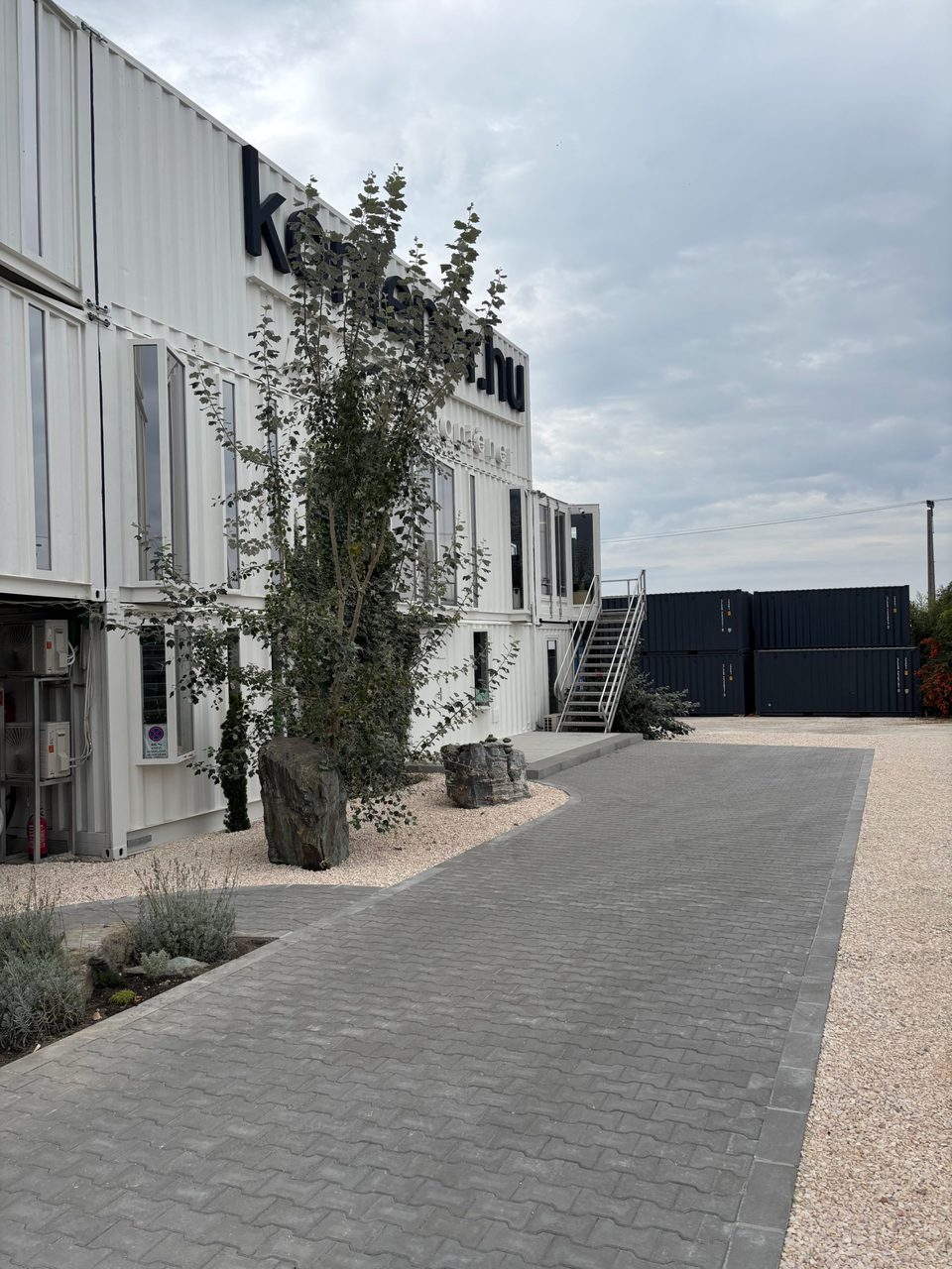 Az érdi irodakonténer bemutató telephely: emeletes konténeriroda-épület kontener.hu felirattal, előtte térkövezett bejáró, háttérben tengeri konténerek